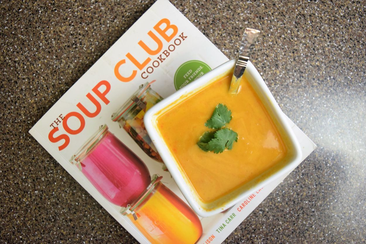 square bowl of orange soup sitting on the Soup Club Cookbook