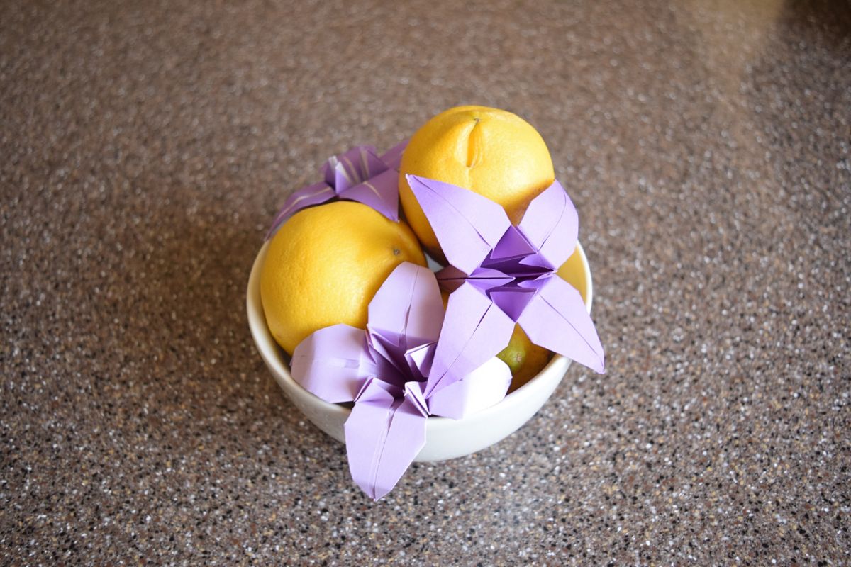 3 purple origami lilies in a white bowl with 2 yellow lemons on a gray speckled countertop